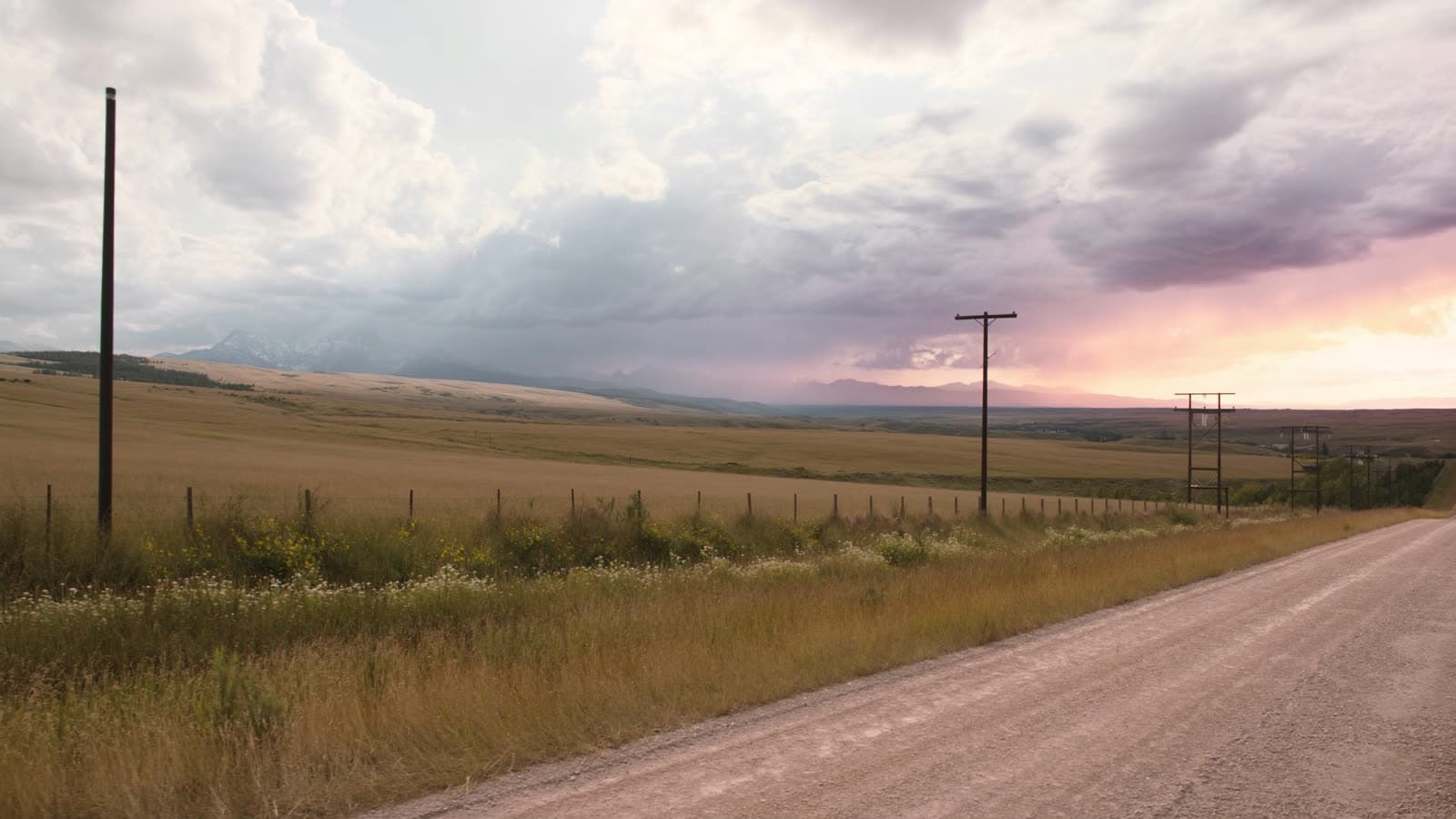 Gravel road and fenced grassland under a storm-lit Montana sky